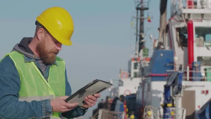 戴着黄色头盔的胡须的港口工人站在港口的平板电脑上，以船为背景。码头工人检查工业港口。电影4k视频