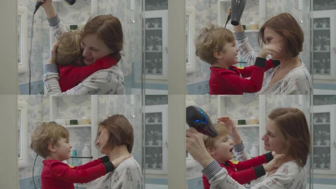 年轻的母亲用吹风机吹干儿子的头发。穿着红色睡衣的孩子在吹干头发时拥抱母亲。有趣的家庭沐浴程序。母子俩