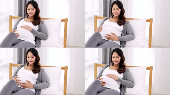 快乐的亚洲孕妇坐在家里卧室的床上，看着她裸露的肚子，怀孕，人和产妇的概念
