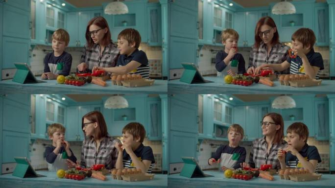一家人一起烹饪站在蓝色厨房的平板电脑上看在线食谱。两个孩子在家帮妈妈做饭。孩子们吃甜红辣椒。