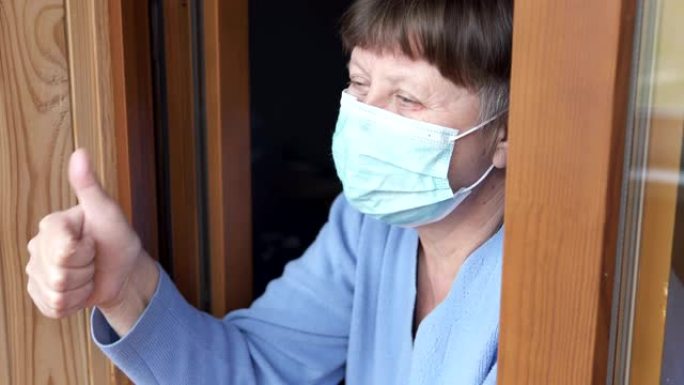 一位戴着医用口罩的老妇看着窗外，挥舞着她的手。检疫自我隔离，冠状病毒，covid 19。
