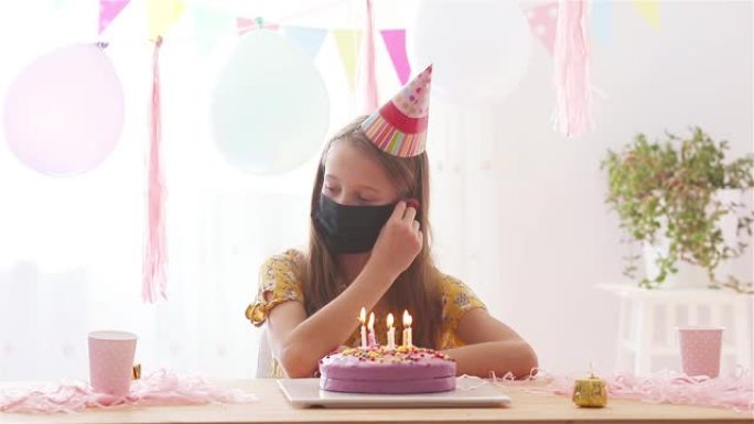 悲伤的小女孩在她的生日聚会上戴口罩以防止病毒。带有气球的节日色彩缤纷的背景。独自过生日。呆在家里检疫