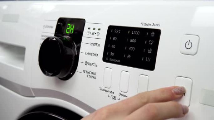 一名妇女转动操纵杆，选择洗衣机上的模式并将其打开。翻译: “棉花，合成材料，羊毛，快洗，冷，温度，拧