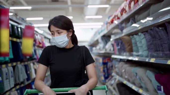 亚洲妇女戴着口罩，在超市购物中心推着购物车慢动作，新型冠状病毒肺炎大流行。年轻女士储备物资，反检疫和