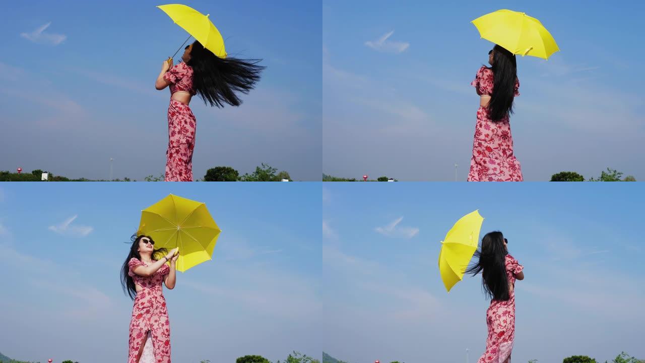戴着墨镜的快乐亚洲女人在蓝天下撑着黄色的伞。