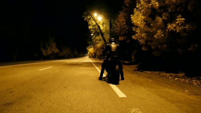 戴着头盔和皮夹克的骑自行车的人来到他的摩托车上，坐在上面，开始在夜间高速公路上骑行。男人在晚上开始骑