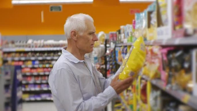 超市里的高级男人。穿着蓝色衬衫的白发老人选择零食，并将食物篮放在商店货架背景上。