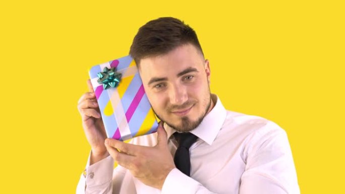 一个穿着衬衫和领带的男人在黄色背景上摇动一个带有礼物的盒子。