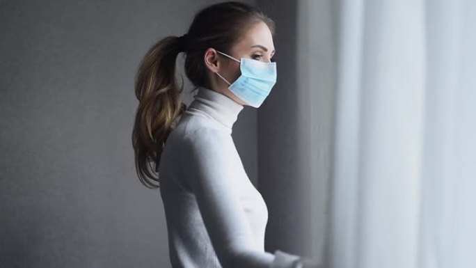 大流行冠状病毒。世界各地的Covid 19。一个戴着一次性医用口罩的年轻女子看着窗外。检疫生活和自我
