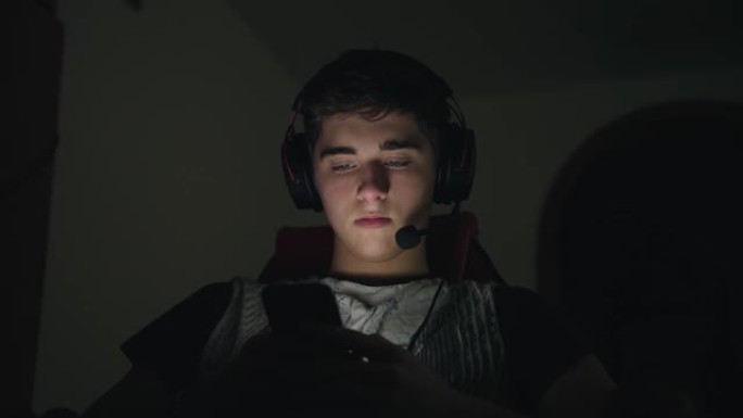 年轻的帅哥戴着带麦克风的耳机，使用智能手机，深夜坐在家里的电脑附近。带智能手机的发光脸