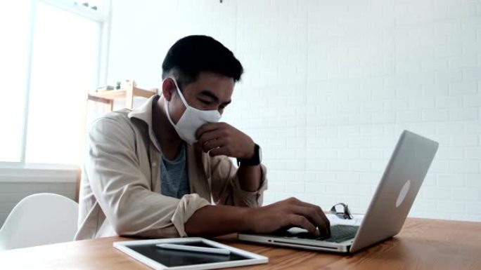 亚洲男子坐在办公桌前，打喷嚏护卫，在家庭办公室使用笔记本电脑，戴着防护口罩，在新型冠状病毒肺炎大流行