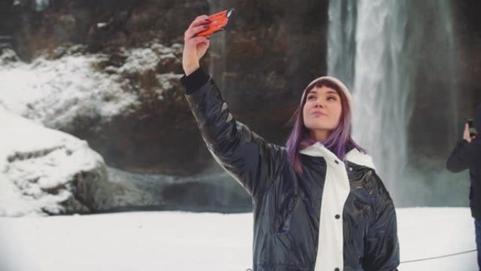 美丽的年轻女子在冰岛的一个瀑布附近，在电话上拍照，欢欣鼓舞并自拍