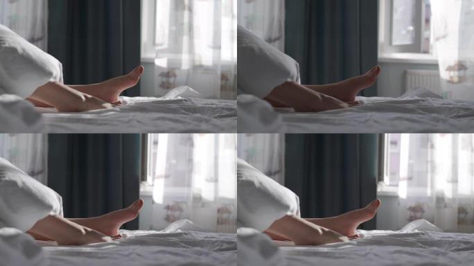 睡在床上的女人盖着白色的毯子，美丽的光脚被阳光照亮