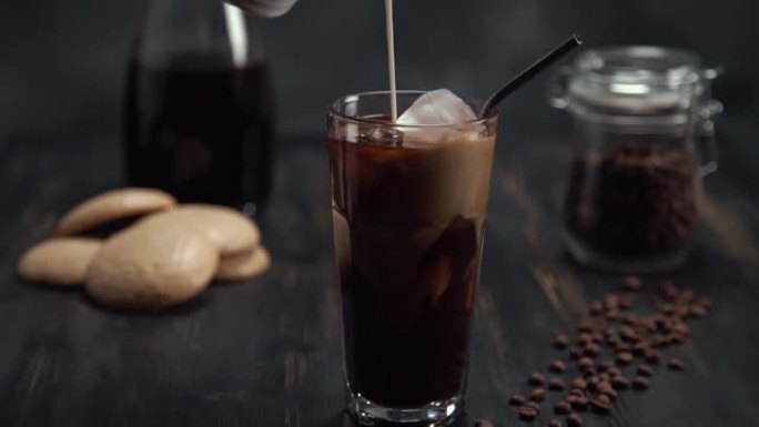 将奶油慢动作倒入黑木桌上的高杯冷煮冰咖啡中，黑色背景上有咖啡豆和饼干