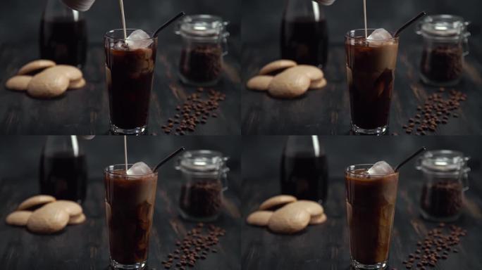 将奶油慢动作倒入黑木桌上的高杯冷煮冰咖啡中，黑色背景上有咖啡豆和饼干