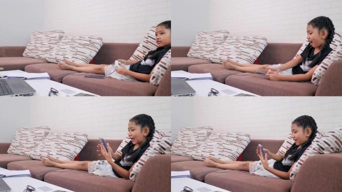 亚洲小姑娘在家里的沙发上玩游戏和妈妈接视频通话。