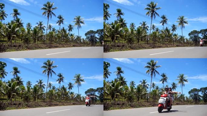 幸福的夫妇在棕榈树林立的道路上行驶时，伸出双臂驾驶轻便摩托车。年轻的男女游客在热带岛屿上沿着沥青车道