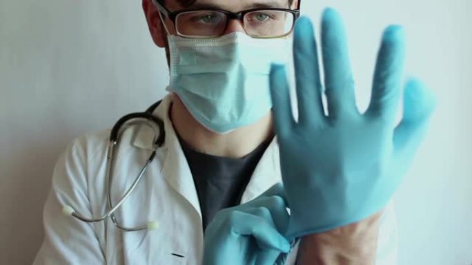 戴着眼镜和医用口罩的年轻英俊医生戴上无菌手套，为患者的检查做准备。一位年轻医生正在准备病人检查。