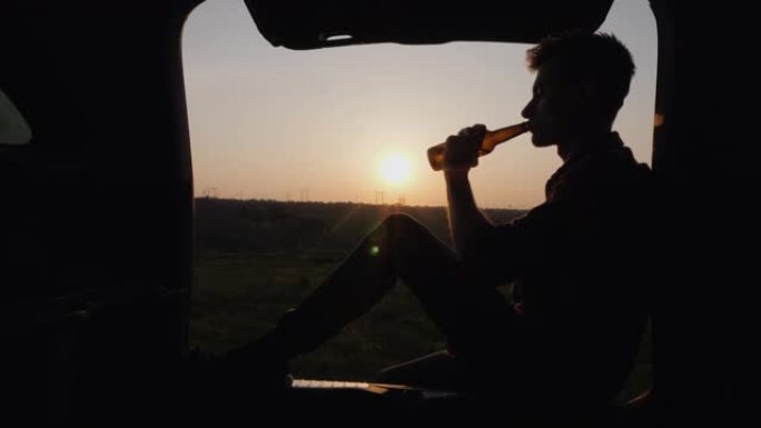 一名少年坐在汽车后备箱里，从瓶子里喝啤酒