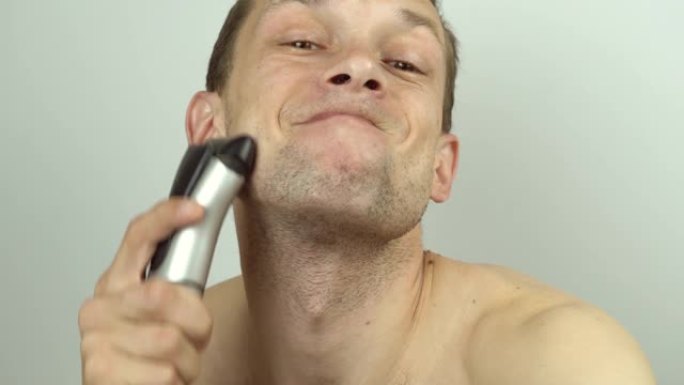一个年轻英俊的男人用电动剃须刀刮胡须，在镜头前胡闹，做着可笑的鬼脸。