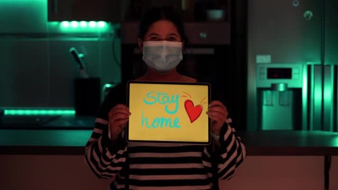 在冠状病毒covid 19卫生危机期间，戴着外科口罩的可爱的微笑女孩在平板电脑上分享 “呆在家里” 