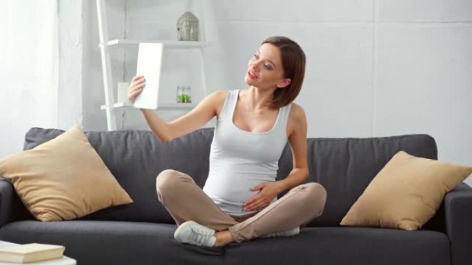 快乐怀孕的女孩在沙发上的数字平板电脑上进行视频通话