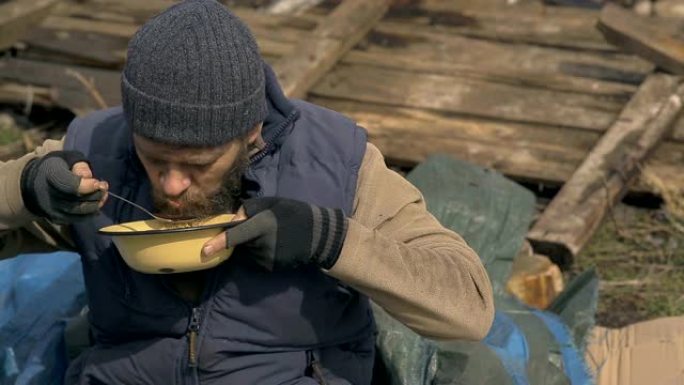 一个无家可归的人从废墟附近的盘子里吃汤，在疫情期间帮助贫困和饥饿的人