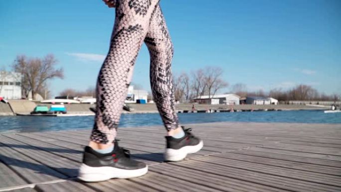 一个年轻的运动女孩穿着黑色运动鞋和绑腿在湖边的码头上奔跑。慢动作。特写。