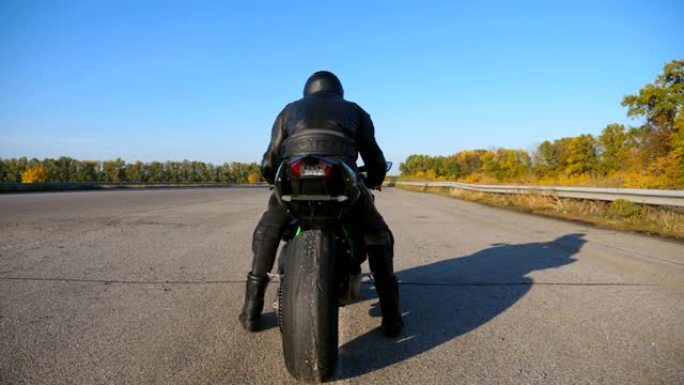 身穿头盔和皮夹克的无法识别的摩托车手的后视图正坐在高速公路上的摩托车上，准备骑行。男人要骑摩托车。自