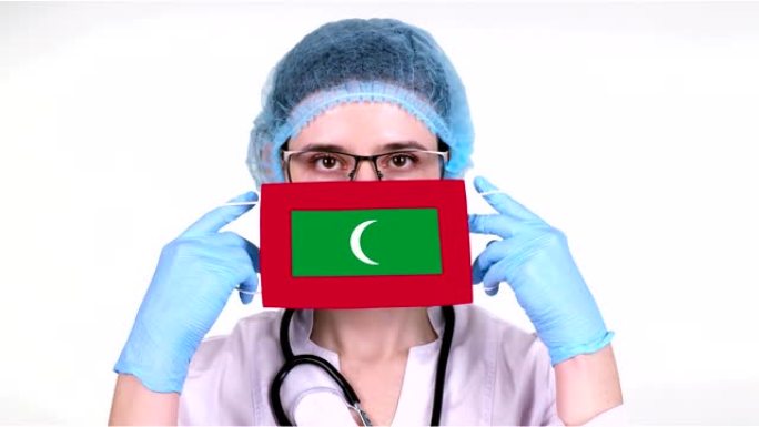 特写。戴眼镜的医生，蓝色医疗帽，手套手持带有马尔代夫国旗的医用口罩。医生护理，冠状病毒期间国家保护，