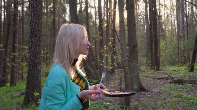 年轻漂亮的金发女郎在森林里野餐。她是一名游客。她手里拿着一个平底锅。
