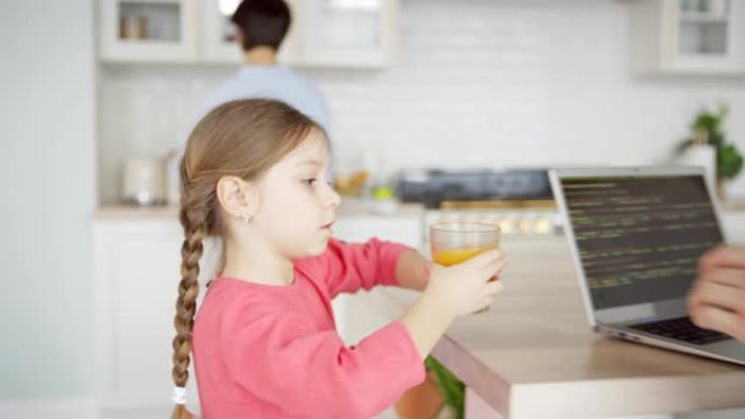 跟踪小女孩在厨房里帮助母亲并为忙碌的爸爸在家里的笔记本电脑上编码的果汁，父亲亲吻乐于助人的女儿的镜头