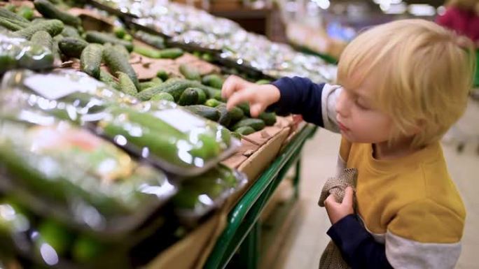 可爱的小男孩选择新鲜的有机黄瓜，然后放入食品店或超市的网状购物袋中。有孩子的年轻家庭的健康生活方式。