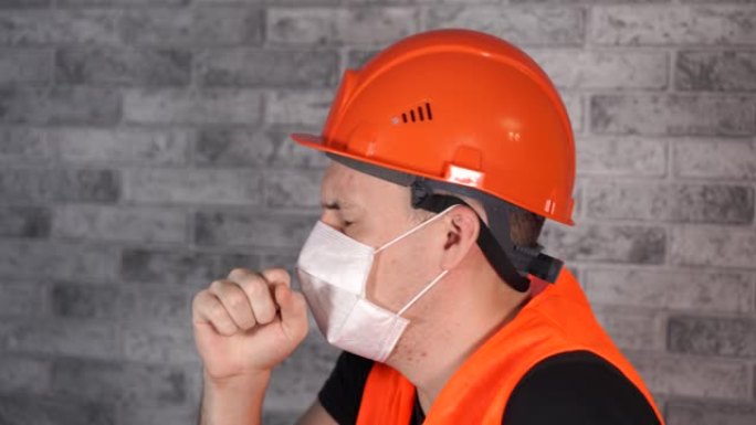 穿着工作服和医用口罩的男性建筑工人在灰色砖墙的面部背景上咳嗽。感染威胁的概念