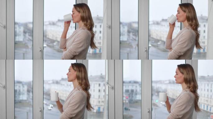 沉思美丽的金发女人喝茶看着窗户。用4K红色相机拍摄