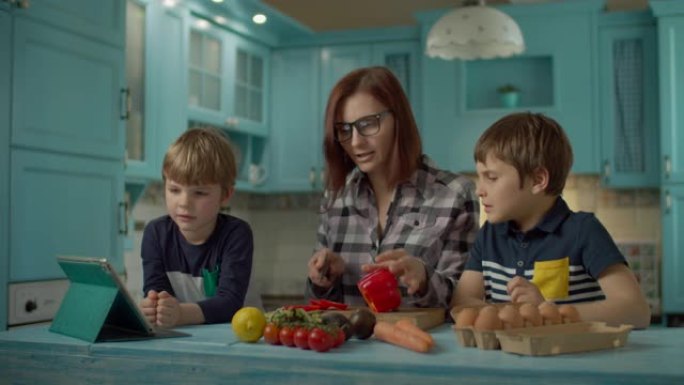 一家人一起烹饪站在蓝色厨房的平板电脑上看在线食谱。两个孩子在家帮妈妈做饭。女人切甜红辣椒。