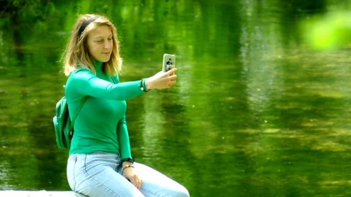 美丽的年轻女子在森林河上的智能手机上拍照