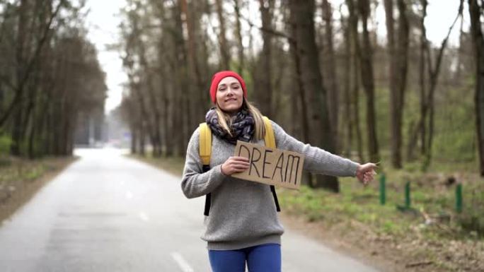 等待的车。年轻的搭便车女人带着海报梦想上路。手指向上