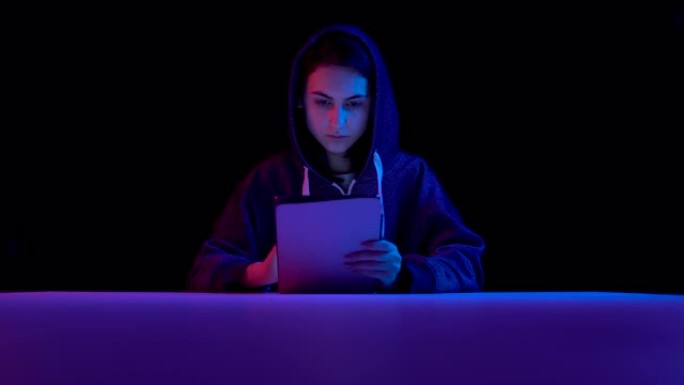 年轻女子头罩着平板电脑。黑客通过平板电脑进行黑客攻击。蓝色和红色的光落在黑色背景上的女人身上。