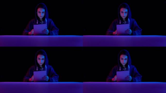 年轻女子头罩着平板电脑。黑客通过平板电脑进行黑客攻击。蓝色和红色的光落在黑色背景上的女人身上。