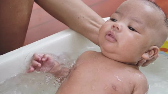 一位亚洲母亲在白色塑料儿童浴缸的浴室里为男婴洗澡的照片