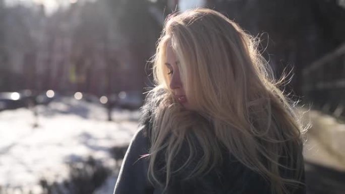 美丽的金发微笑的年轻女子在外面享受冬天的雪和阳光明媚的日子看着相机，微笑，散步的肖像