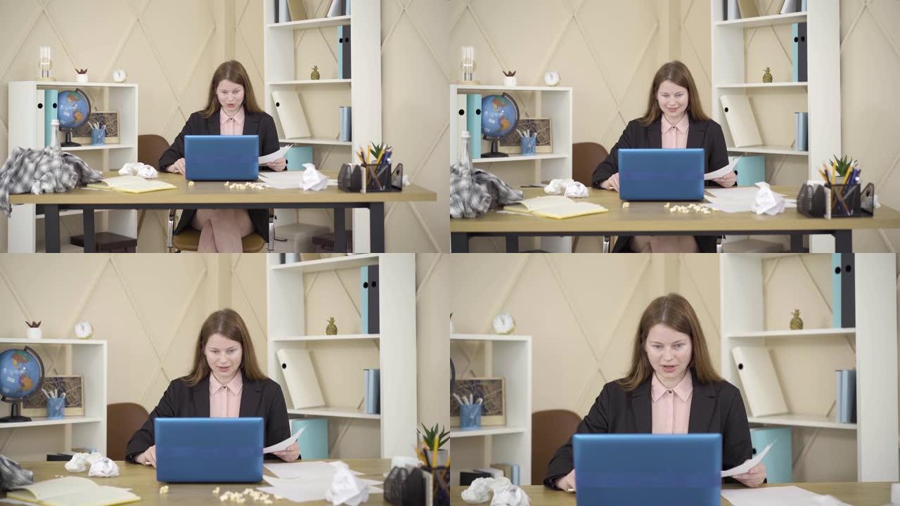 相机接近微笑的年轻红发女人，她坐在笔记本电脑前凌乱的房间里聊天。白人自由职业者在家工作的肖像。生活方