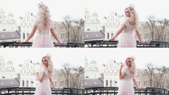 美丽的年轻性感女人站在阳台上，摆姿势背景冬春老城市景观。金发公主可爱的脸优雅的连衣裙。健康白色长卷卷