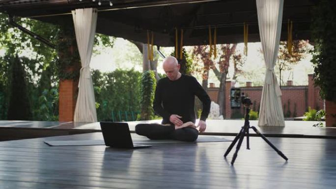 穿着黑色运动服的瑜伽教练正坐在莲花的位置，看着笔记本电脑。用智能手机在庭院的三脚架上拍摄自己的视频