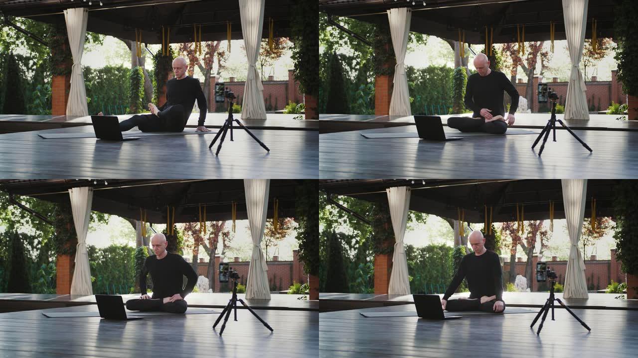 穿着黑色运动服的瑜伽教练正坐在莲花的位置，看着笔记本电脑。用智能手机在庭院的三脚架上拍摄自己的视频