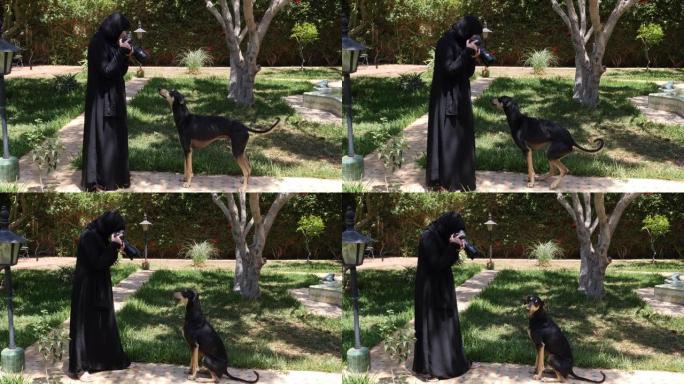 一位美丽的摩洛哥阿拉伯穆斯林妇女拿着数码单反相机在花园里给一只黑色的Sloughi狗(阿拉伯灰狗)拍