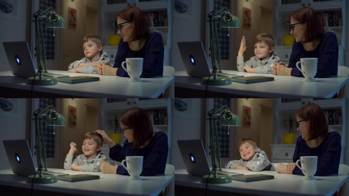 30多岁的年轻母亲在家与学龄前儿童的儿子一起做在线作业。在网上学习时，小男孩站起来到笔记本电脑屏幕上
