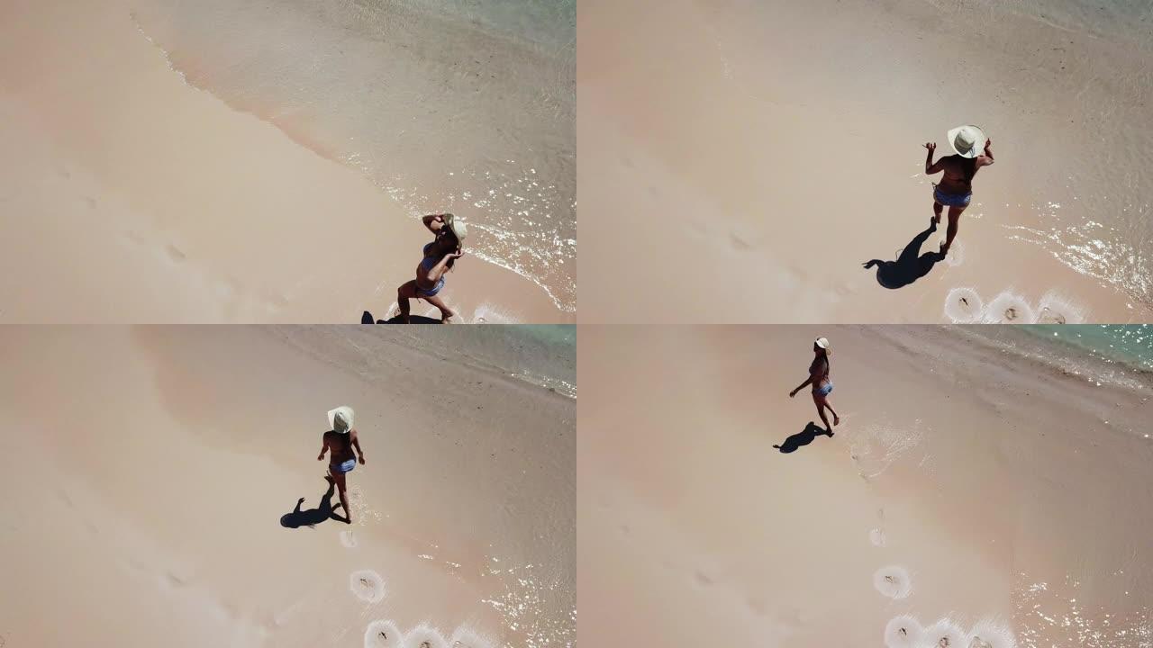 在印度尼西亚龙目岛粉红海滩的海边，一个戴着草帽的女孩在沙滩上行走的特写无人机镜头。波浪轻轻地冲刷着海