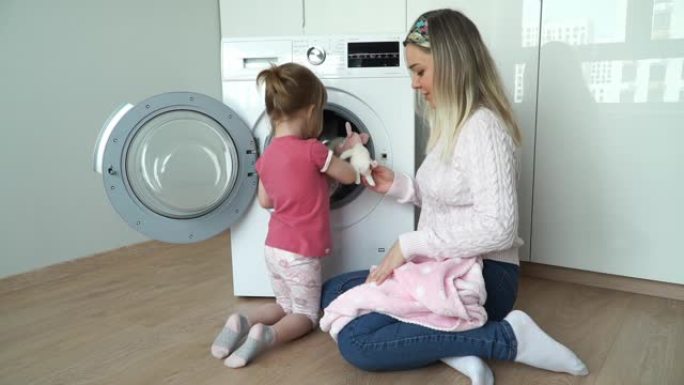 小微笑的3岁女儿的肖像和年轻的母亲把柔软的玩具放在洗衣机里洗。女孩关闭舱门，打开家用电器。